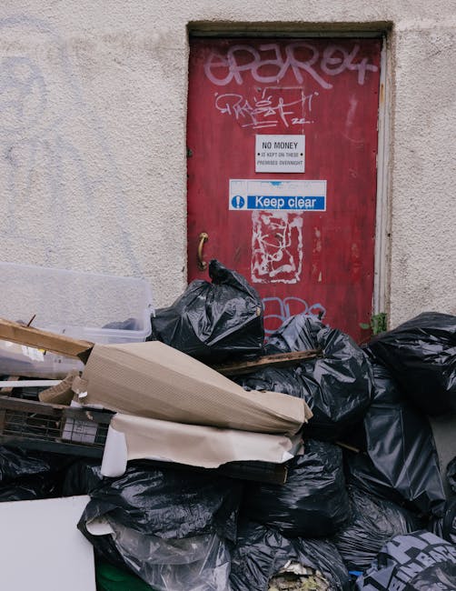 An outdoor scene featuring a weathered red metal door with graffiti tags, stickers, and warning signs, including a 'No Money' notice and a 'Keep Clear' sticker, set in a rough, textured beige wall. In front of the door, there is a pile of black trash bags filled with discarded items, some partially open revealing packaging and debris. To the left, a wooden furniture piece and cardboard packaging are propped against the bags, with plastic wrapping and protective padding visible among the discarded materials. The area appears to be on a pavement or alleyway, and the overall scene suggests a packaging or disposal context related to house removals or transporting belongings during a move, as handled by Man with Van The Burroughs who specialise in services such as furniture transport and home relocation. The lighting is natural, indicating daytime, emphasizing the cluttered state typical of moving or clearance activities outside residential or commercial premises.