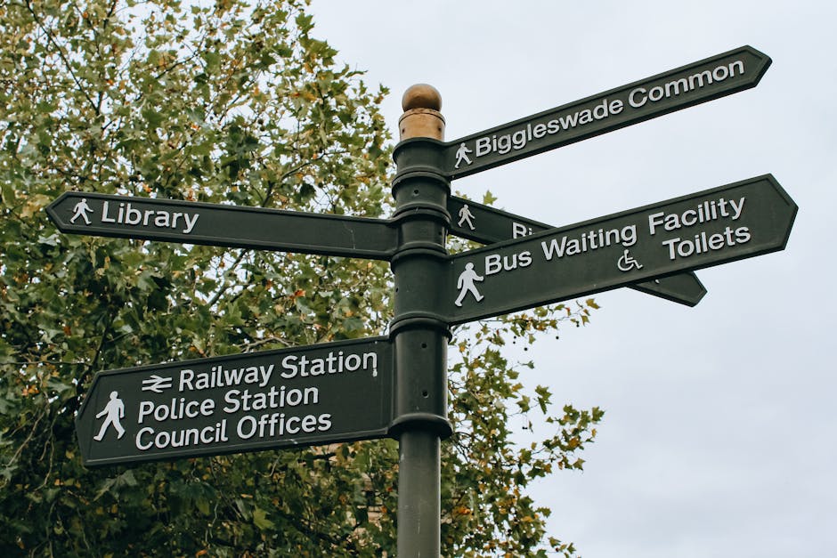 A black multi-directional street signpost situated outdoors with six rectangular signs pointing in various directions, each displaying white text and icons indicating nearby locations. The signs indicate directions to Biggleswade Common, Library, Railway Station, Police Station, Council Offices, Bus Waiting Facility, and Toilets. The signpost is mounted on a post with a rounded top, set against a background of leafy trees and a cloudy sky. The surrounding environment suggests a public area that could be near residential or community buildings. This setting reflects a typical outdoor navigation aid that residents or visitors in NW4 might see when orienting themselves for local amenities and transport links, relevant to house removals or outdoor logistics associated with relocations. The scene is lit by natural daylight, with visible greenery and foliage, contributing to a clear and informative street or community signposting context.