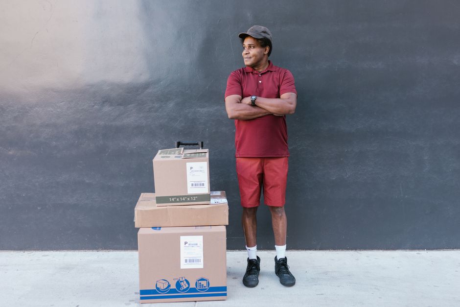 A man wearing a grey cap, red polo shirt, red shorts, white socks, and black sneakers stands outdoors against a dark grey wall with his arms crossed, smiling and observing a loading process. In front 
