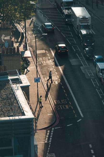 An aerial view of a busy city street during daylight showcasing a parking area with marked bus stops on the pavement and multiple vehicles, including cars and vans, waiting or moving along the road. Two individuals, one dressed in a high-visibility vest and the other in casual clothing, are positioned on the sidewalk; the one in high-visibility appears to be managing traffic or assisting with a home relocation process, while the other is walking nearby. The scene includes nearby trees providing partial shade, with shadows cast across the pavement indicating a sunny day. This setting captures a typical transport and moving environment where furniture and packing materials from house removals by Man with Van The Burroughs may be prepared for transport, reflecting the logistical aspect of packing and loading during a house move from Brent Cross to The Burroughs.