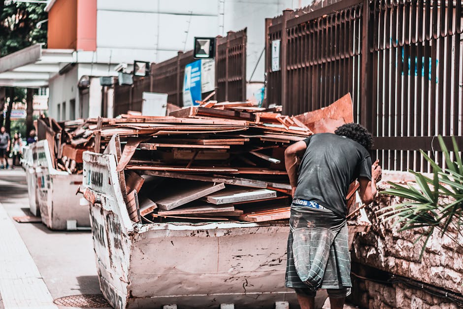 A man with curly hair wearing a black t-shirt and patterned shorts is standing beside a large, overflowing skip filled with a variety of waste materials including wooden panels, cardboard, and plastic wrapping. The skip is placed on a pavement in an urban environment next to a high, brown metal fence, with a building visible in the background. Traffic signs and other urban fixtures can be seen along the sidewalk. The man appears to be examining or preparing to load or unload items as part of a home relocation or clearance process. This scene illustrates a typical bulky waste removal associated with house removals or professional furniture transport services offered by Man with Van The Burroughs, highlighting the logistics involved in packing, handling, and disposing of large waste during a furniture removal or moving project.
