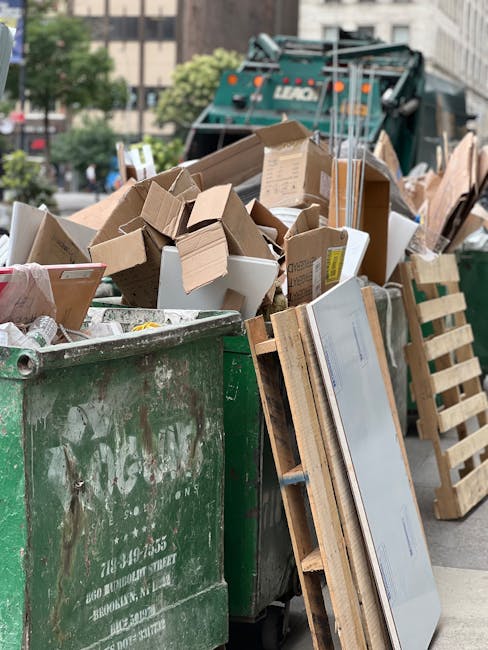 A large open green waste skip filled with various bulky waste items, including cardboard boxes, wooden pallets, packaging materials, and flattened furniture parts, situated outdoors on a city street curb. In the background, a commercial waste collection truck is visible, parked behind the skip, with trees and tall buildings lining the street. The scene depicts the collection and disposal process of household refuse, relevant to home relocation or moving services provided by Man with Van The Burroughs, illustrating the handling of bulky waste during house removals or packing and moving operations.