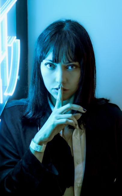 A woman with shoulder-length dark hair and light skin is standing indoors near a window with bright natural light. She is holding her index finger vertically against her lips in a gesture of silence. She is wearing a dark jacket over a light-colored shirt, with a watch on her left wrist. The background features a blurred wall with some illuminated signage or window light, creating a professional yet casual atmosphere. The image emphasizes a quiet, discreet approach, suitable for illustrating concepts related to confidential home relocation, packing, or moving services by Man with Van The Burroughs, with a focus on careful handling and unobtrusive moving processes.