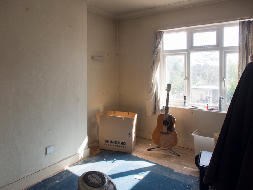 A room during a home relocation process shows a large window with partially drawn curtains letting in daylight, illuminating the space. Near the window, an acoustic guitar with a wooden finish rests on a stand, positioned on a wooden floor with a small, dark blue rug. sunlight creates shadows on the floor. Against the wall, there is an open cardboard box labeled 'SHURGARD SELF-STORAGE' containing packing materials. A white plastic storage bin is situated next to the window, partially visible. To the right, a black garment or cover is hanging, and a small vacuum cleaner is on the floor near the doorway. The room appears to be in the midst of packing or preparation for furniture transport, with loose items and minimal furnishings, typical in a house moving or packing scenario, as managed by companies like Man with Van The Burroughs.