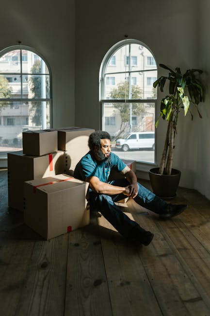 A person with curly hair and medium skin tone, dressed in a blue shirt and dark trousers, sitting on a wooden floor next to several cardboard moving boxes wrapped with red tape. The boxes are stacked behind the individual, some upright and others lying flat. The room features two large arched windows allowing natural light to illuminate the scene, with a view of parked cars and trees outside. A tall potted plant is situated near the windows on the right side, adding greenery to the space. This setting appears to be part of a home undergoing a move or packing process, with the individual resting during the home relocation or packing and moving activities carried out by [COMPANY_NAME], a professional removals service. The scene captures an interior space during a furniture transport or loading process, emphasizing the careful handling of boxes and the preparation for a house transfer.