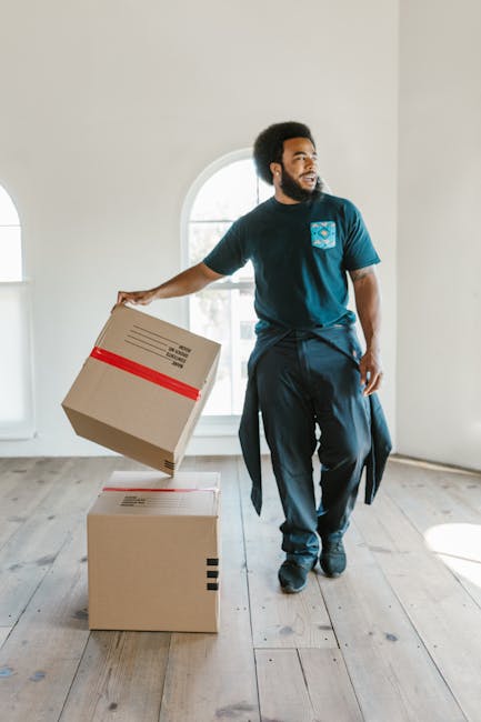 A man with dark skin and curly black hair, dressed in a turquoise T-shirt with a logo on the chest and dark trousers, is shown inside a bright, empty room with wooden flooring and white walls. He is holding a cardboard box with red tape around it in his right hand, preparing for a furniture transport or home relocation. Two additional cardboard boxes are positioned on the floor nearby, one on top of the other, possibly filled with household items. The room features a large, arched window that allows natural light to illuminate the space, indicating it is daytime. The scene captures the packing and moving process, aligning with professional removals services provided by Man with Van The Burroughs, focusing on efficient packing and furniture transport during house removals.