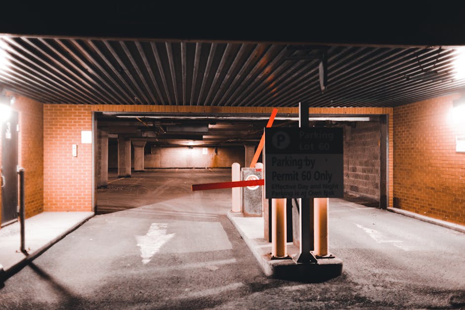 A dimly lit underground parking area with brick walls and a ceiling made of dark, parallel metal slats. In the foreground, a metal barrier arm is lowered across the entrance, attached to a sign indicating parking restrictions and permit requirements. The pavement features white directional arrows and is surrounded by concrete curbs. To the left, a brick wall has a small keypad or access panel, and a narrow sidewalk runs along the wall. In the background, the parking garage extends into dimmer, open space with concrete supports and additional parking bays. The overall scene depicts an organized vehicle access point, suitable for home relocation or furniture transport planning, with the environment prepared for parking control during a house move or similar service by Man with Van The Burroughs.