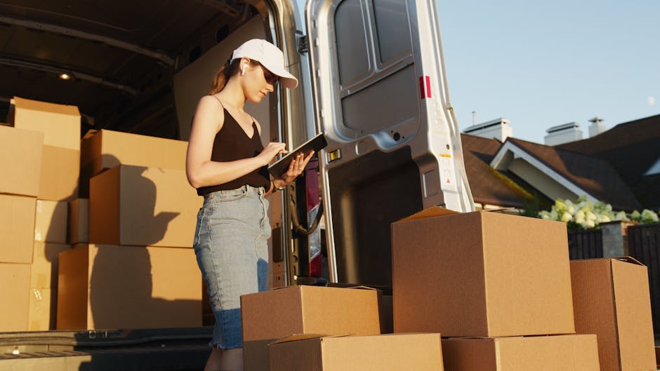 The image depicts the open rear of a white moving van on a paved road, loaded with numerous neatly stacked cardboard boxes of various sizes, prepared for a home relocation. Two individuals are present