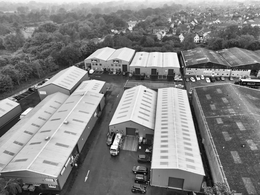 An aerial view in black and white showing a collection of warehouse-style industrial buildings with curved metal roofs, situated in an area with numerous parked cars and trucks around the buildings. I