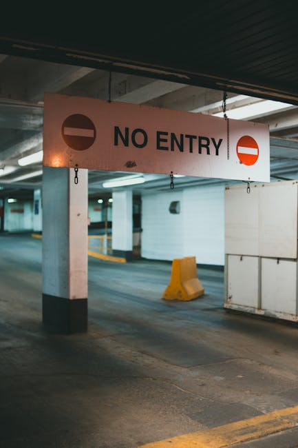 Inside a multi-storey car park or underground parking facility, a 'NO ENTRY' sign with a white background and black text is prominently displayed, accompanied by a red circle with a white horizontal bar indicating the restriction. The sign hangs from a dark, ribbed ceiling and is mounted on a light grey concrete pillar supported by black and white painted sections at the bottom. In the background, the parking area features white walls, concrete floors, and a few yellow wheel stops or bumper blocks positioned near parking spaces. The environment is well-lit with overhead fluorescent lighting, and the overall setting appears clean and organized. This scene is relevant to house removals and moving services, illustrating a typical parking or loading area where furniture transport and packing and moving processes might take place, possibly operated by a professional removals company like Man with Van The Burroughs working within NW4 postal code areas.