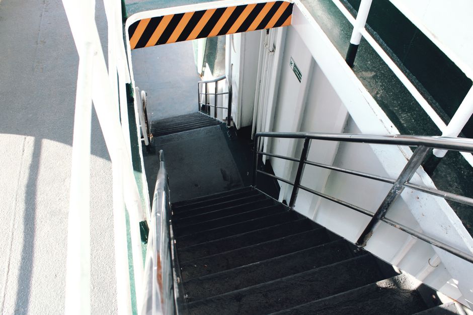 A set of narrow, steep outdoor metal stairs with black steps and silver handrails, leading down from a building's exit door. The exterior walls are white with black and orange striped caution tape along the upper edge of the doorway. The stairs are situated on a paved surface, with sunlight casting shadows on parts of the steps and the surrounding ground. The image captures a moment during a home relocation process, illustrating the challenges of moving large furniture or boxes through tight access points, possibly managed by Man with Van The Burroughs as part of their removals service. The environment is outdoors, adjacent to the building, with a focus on the physical constraints of narrow staircases for furniture transport and packing and moving logistics.