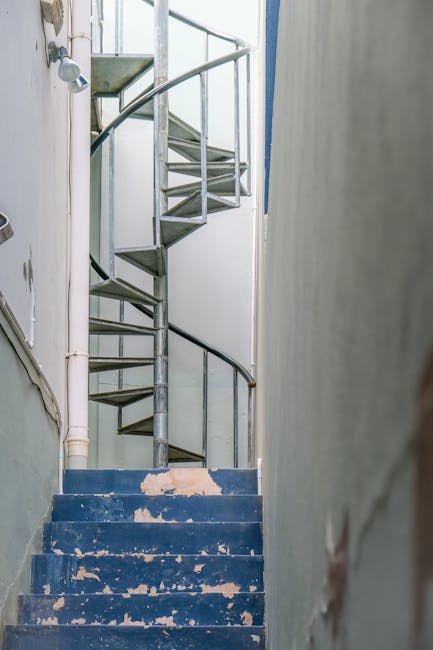 The image depicts a narrow indoor staircase with worn, blue-painted steps showing peeling paint and signs of use. The staircase has a metal handrail on the right side and a spiral metal staircase with perforated steps and a curved handrail connecting to the upper floor, visible at the top. The walls surrounding the staircase are plain, with the left wall featuring a vertical white pipe running alongside the stairs. Lighting appears natural, illuminating the space evenly. This setup illustrates a typical interior of a residential property requiring careful navigation during a home relocation or furniture transport process. Man with Van The Burroughs, a professional removal service, may encounter such narrow-access challenges when planning efficient packing and moving routines within properties like this, especially when managing tight stairs or limited space for furniture manoeuvres.