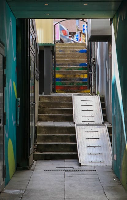A set of narrow, steep outdoor metal stairs with black steps and silver handrails, leading down from a building's exit door. The exterior walls are white with black and orange striped caution tape along the upper edge of the doorway. The stairs are situated on a paved surface, with sunlight casting shadows on parts of the steps and the surrounding ground. The image captures a moment during a home relocation process, illustrating the challenges of moving large furniture or boxes through tight access points, possibly managed by Man with Van The Burroughs as part of their removals service. The environment is outdoors, adjacent to the building, with a focus on the physical constraints of narrow staircases for furniture transport and packing and moving logistics.