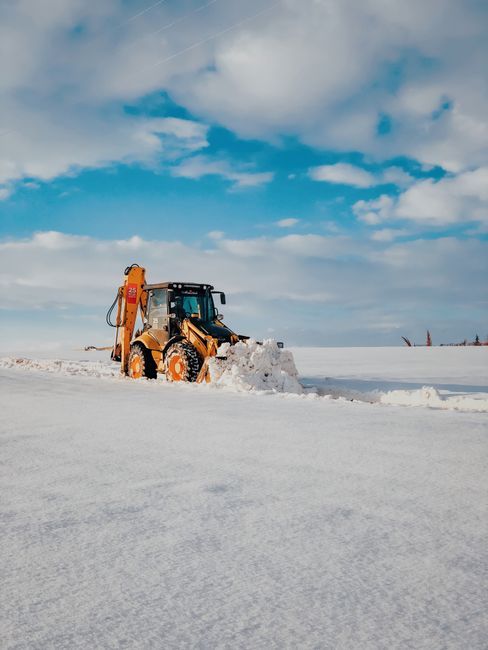 A yellow front-end loader equipped with a large bucket attachment is clearing snow on a flat, snow-covered landscape under a partly cloudy sky. The machine is actively scooping and pushing snow, creating a pile in front of its bucket. The surrounding area is vast and open, with no other structures or vehicles visible, indicating an outdoor environment suitable for heavy-duty snow removal or land clearing. Images of equipment like this are relevant to professional removals and transportation services, especially in regions prone to snow. Man with Van The Burroughs offers such services including handling heavy items and home relocations, which may involve outdoor logistics and equipment use during winter months.