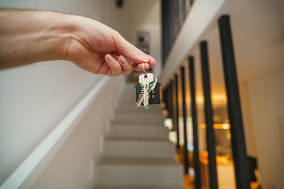 A close-up of a person's hand holding a set of house keys with multiple keys on a keyring, positioned above a staircase inside a building. The staircase has white risers and dark handrails with vertical black balusters, leading upward into a well-lit interior space. The background shows a softly illuminated corridor with warm lighting, suggesting an environment prepared for home relocation or moving activities. This scene reflects the process of moving out or evictions, with the keys symbolising access and handover during a furniture transport or house clearance service. The image is associated with professional removals, as provided by Man with Van The Burroughs, who specialise in rapid clearance and relocation services in NW4.