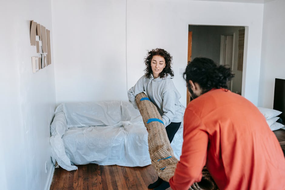 A woman with curly dark hair, dressed in a light grey hoodie, is assisting a man with dark, curly hair and a beard, wearing an orange jacket, in carrying a large rolled-up piece of furniture wrapped in brown fabric with blue straps. They are inside a room that has a white wall with a small decorative art piece, and a white couch covered with a plastic sheet. The man is holding the furniture piece while the woman supports it from the side, engaged in a home relocation and furniture transport process. The flooring is wooden, and there is a doorway in the background leading to another room. This scene illustrates a packing and moving activity, with the team working together to load furniture for a house move, possibly as part of a professional removals service by Man with Van The Burroughs, focusing on rapid clearance for eviction or urgent landlord moves in NW4.