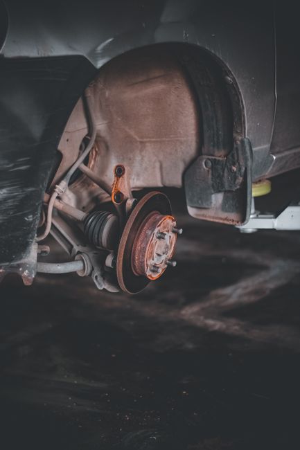 Close-up of a car's front wheel arch with the wheel removed, exposing the brake disc, suspension components, and inner wheel well. The vehicle is parked on a dark surface, and the surrounding area is dimly lit, emphasizing the mechanical parts. This image illustrates a vehicle undergoing maintenance or preparation for a home relocation involving furniture transport or loading. Man with Van The Burroughs offers removals and moving services, supporting efficient loading processes during house moves. The focus on the vehicle's exposed brake disc and suspension highlights the importance of proper vehicle maintenance in safe transportation of furniture and household items during a home relocation or packing and moving process.