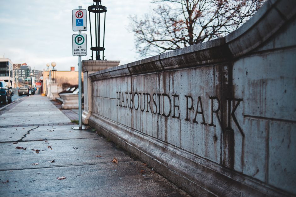 A close-up view of a stone wall with the engraved inscription 'HARBOR SIDE PARK' along the sidewalk of The Burroughs in an urban area. The pavement in the foreground has some scattered leaves and cracks, indicating outdoor conditions. Behind the wall, street signs indicate parking regulations, including a disabled parking space and a general parking zone with time restrictions from 8 am to 6 pm. To the left, parked cars line the street, and in the background, buildings and streetlights are visible under an overcast sky, suggesting a typical city environment suitable for home relocation or furniture transport activities. The presence of street signs and the urban setting support the context of moving logistics, possibly involving vehicle access or parking suspensions for house removals managed by Man with Van The Burroughs.